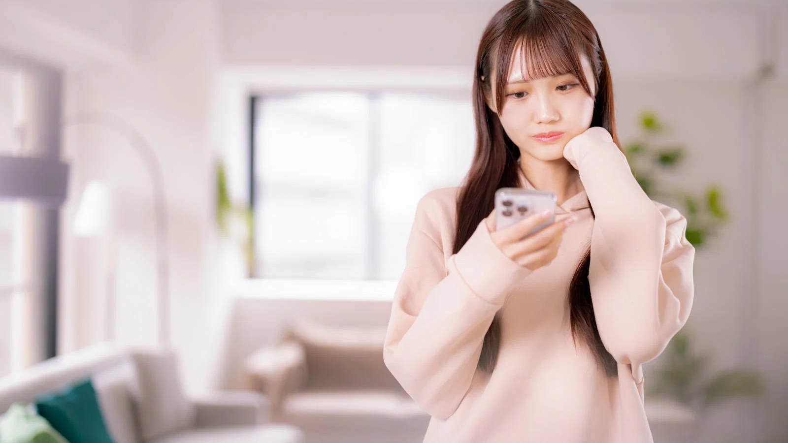 明るい部屋でスマートフォンを見ながら考える女性