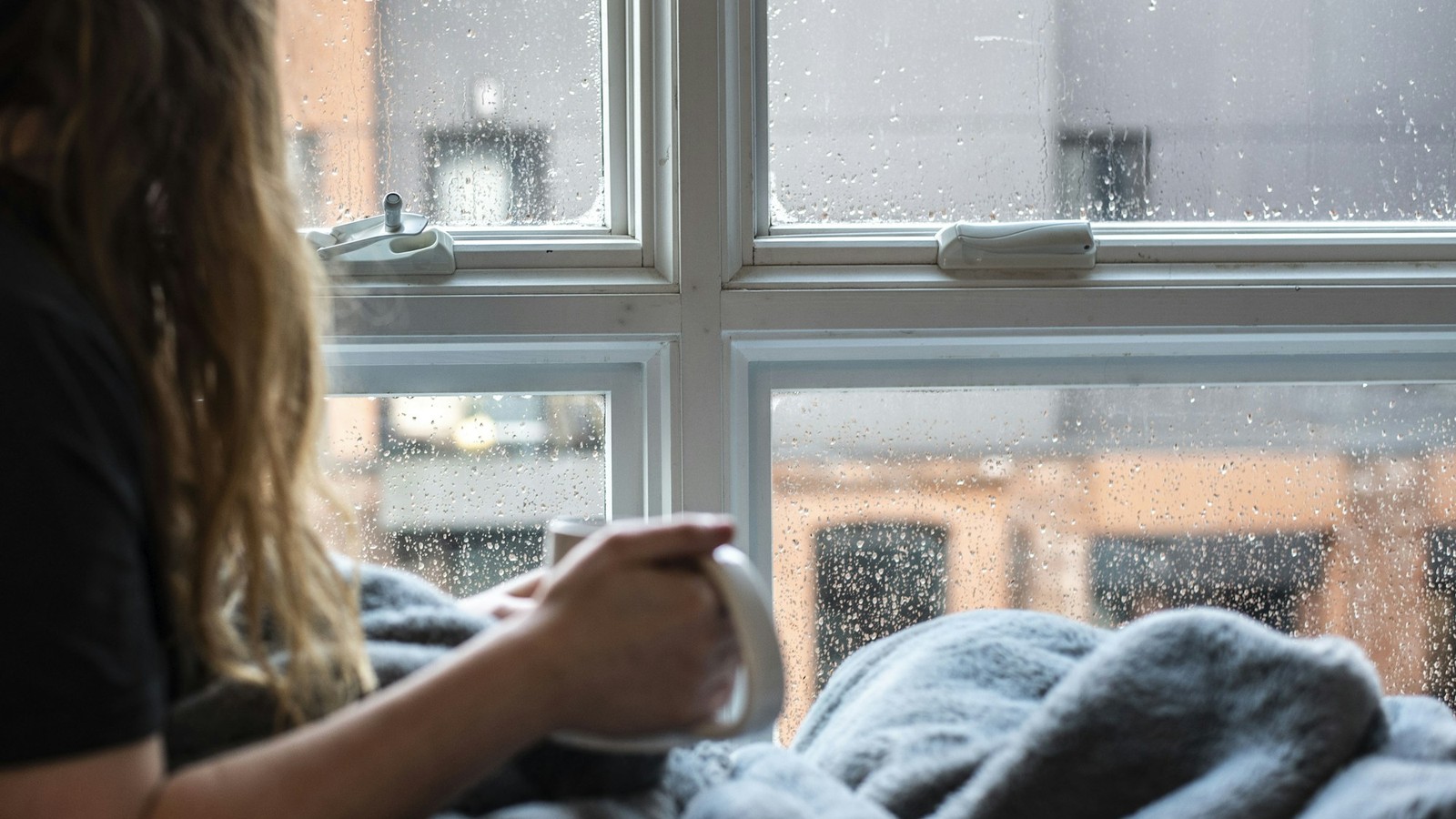 雨の窓辺でマグカップを両手で持つ女性の手元